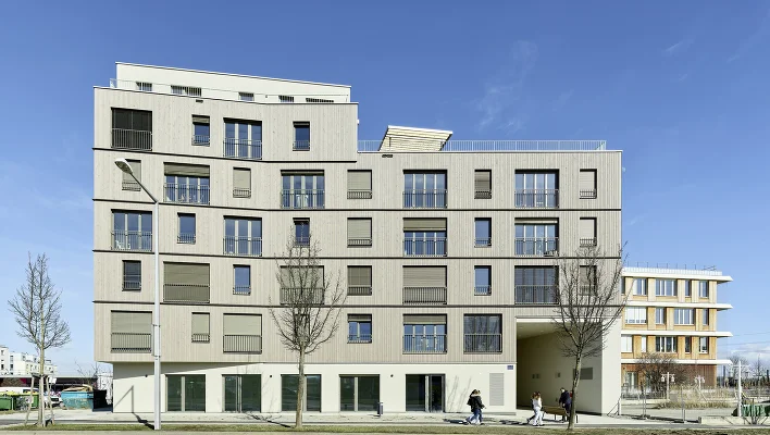 Wohngebäude mit Holzfassade an einer Straße, davor Gehweg mit Bäumen und vorbeigehenden Personen.