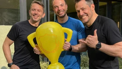 Drei  Männer in Sportkleidung vor einem gelben Gebäude mit Plastikpokal